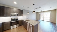 This thoughtfully designed one-bedroom, one-bathroom unit in Round Rock, Texas offers an efficient and modern living space across a single floor. The open-concept layout maximizes functionality, with the kitchen serving as a central highlight. Dark espresso cabinetry extends to the ceiling, providing ample storage, while granite countertops and a classic white subway tile backsplash add a polished aesthetic. A peninsula island with an undermount sink and pendant lighting above creates a natural gathering point and flows seamlessly into the living area. Stainless steel appliances, including a French door refrigerator, electric range, built-in microwave, and dishwasher, complete the kitchen. Light wood-tone flooring runs throughout the main living areas, adding warmth to the space. The living area connects to a private balcony through sliding glass doors, allowing natural light to fill the room. The bedroom features soft carpet flooring, a ceiling fan, and a window with exterior views. It connects directly to the full bathroom, which includes a glass-enclosed walk-in shower with large-format tile surround. Adjacent to the bathroom is a generously sized walk-in closet outfitted with built-in shelving, hanging rods, and corner shelving units for organized storage.