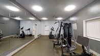 This fitness center offers a practical and functional workout environment designed to support a range of exercise routines. The space is finished with durable, speckled rubber flooring ideal for high-traffic athletic use, and the walls are painted in a clean, neutral gray tone that contributes to a focused atmosphere. Large wall-to-wall mirrors line one side of the room, visually expanding the space and allowing users to monitor their form during exercise. Overhead, flush-mounted ceiling lights provide bright, even illumination throughout the room, complemented by a textured ceiling finish. The facility is equipped with key cardio machines, including a treadmill and an elliptical trainer, positioned to allow comfortable use. A prominent Omega multi-station strength training machine serves as a centerpiece, offering a variety of resistance-based exercise options. An exercise chart posted on the wall provides guidance for users. The space also includes doorway access to additional areas, such as restrooms or utility rooms, enhancing overall convenience. The layout is efficient and well-organized, making the most of the available floor space while keeping equipment accessible. This fitness center is well-suited for residents or members seeking a straightforward, no-frills workout facility.