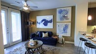 This single-floor space makes a strong design statement through its thoughtfully curated living area. The room is anchored by a deep navy blue tufted chaise sofa, which serves as the focal point and sets the tone for the bold color palette throughout. Warm taupe-toned walls provide a rich backdrop that complements the blue accents seamlessly.

The walls are adorned with striking large-scale abstract artwork — a single oversized piece on one wall and two stacked framed prints on the adjacent wall — all featuring fluid blue brushwork that ties the room's color scheme together cohesively. A ceiling fan with an integrated light fixture provides both airflow and ambient lighting, while a sleek tripod floor lamp adds supplemental task lighting.

Dark, patterned curtains frame the window, adding texture and depth to the space. Hardwood-style flooring runs throughout, grounded by a plush area rug beneath a round metallic coffee table. Decorative accents, including a geometric lantern, small sculptures, and a succulent, add personality and layers of visual interest, resulting in a space that feels both sophisticated and inviting.