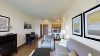 This single-floor unit showcases an open-concept design that seamlessly integrates the living, dining, and kitchen areas into one cohesive space. The living area is anchored by a comfortable grey sofa and matching armchair arranged around a tufted ottoman, creating an inviting seating arrangement. Framed artwork adorns the walls, adding a curated, modern touch to the neutral palette.

Adjacent to the living space, a compact dining area accommodates a dark wood table with upholstered chairs, ideal for everyday meals. The kitchen is positioned at the rear of the open floor plan, featuring warm wood-toned cabinetry, stainless steel appliances, and a breakfast bar with bar stools that serves as both a casual dining and prep space. A pendant light above the bar adds a stylish focal point.

Throughout the unit, light hardwood flooring ties the spaces together while enhancing the sense of openness. The neutral wall tones and recessed and flush-mount lighting contribute to a bright, welcoming ambiance. The layout is designed for efficient use of space while maintaining a comfortable and modern aesthetic.