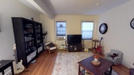 This single-floor space offers a comfortable and functional layout designed for everyday living. The main living area is anchored by rich hardwood flooring that runs throughout, lending warmth and continuity to the room. Two windows fitted with roller blinds allow for adjustable natural light, brightening the space while maintaining privacy. The walls are painted in a soft, neutral cream tone that complements the warm wood tones underfoot.

A large bookcase along one wall provides generous storage for books and personal items, serving as both a functional and decorative element. Seating is arranged thoughtfully around a central area rug, creating an inviting gathering space. A media console positioned between the windows serves as the focal point of the entertainment area. The room also accommodates personal hobbies, with space for a guitar and other personal items that add character and individuality to the space.

Structural beams visible on the ceiling add an architectural detail that gives the room depth and character. The overall ambiance is warm, casual, and personal — a space that balances practicality with the comforts of home, making it well-suited for a variety of daily activities from relaxing to entertaining.