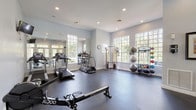 This fitness center offers a comprehensive workout environment designed to accommodate a wide range of exercise routines. The space is housed on a single floor and is characterized by soft gray walls, recessed lighting, and durable rubber flooring throughout, creating a clean and functional atmosphere. Large multi-pane windows line the walls on multiple sides, inviting an abundance of natural light and providing views of the surrounding greenery, giving the space an open and refreshing feel. French doors with transom windows connect to an adjacent outdoor area, further enhancing the sense of openness and accessibility. The cardio section is well-appointed with multiple treadmills, elliptical machines, a Matrix stair climber, and stationary cycling bikes. A dedicated strength training area features a cable pulley machine, a two-tier dumbbell rack with a full set of free weights, along with a rowing bench and functional fitness accessories. A freestanding rack holds medicine balls, resistance bands, and kettlebells, supporting dynamic and functional training. Full-length mirrors along one wall visually expand the space and allow exercisers to monitor their form. Wall-mounted televisions add convenience, while framed artwork provides a welcoming touch, balancing the utilitarian nature of the space with an inviting aesthetic.