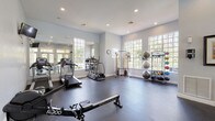 This fitness center offers a comprehensive workout environment designed to accommodate a wide range of exercise routines. The space is housed on a single floor and is characterized by soft gray walls, recessed lighting, and durable rubber flooring throughout, creating a clean and functional atmosphere. Large multi-pane windows line the walls on multiple sides, inviting an abundance of natural light and providing views of the surrounding greenery, giving the space an open and refreshing feel. French doors with transom windows connect to an adjacent outdoor area, further enhancing the sense of openness and accessibility. The cardio section is well-appointed with multiple treadmills, elliptical machines, a Matrix stair climber, and stationary cycling bikes. A dedicated strength training area features a cable pulley machine, a two-tier dumbbell rack with a full set of free weights, along with a rowing bench and functional fitness accessories. A freestanding rack holds medicine balls, resistance bands, and kettlebells, supporting dynamic and functional training. Full-length mirrors along one wall visually expand the space and allow exercisers to monitor their form. Wall-mounted televisions add convenience, while framed artwork provides a welcoming touch, balancing the utilitarian nature of the space with an inviting aesthetic.