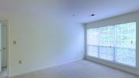 This single-floor residential space offers a clean, minimalist aesthetic defined by neutral tones and an open, airy atmosphere. The interior is finished with soft, wall-to-wall carpeting that adds warmth and comfort underfoot, while smooth white walls and white ceilings contribute to a bright and welcoming environment throughout.

A standout feature of the space is the large window fitted with horizontal blinds, which floods the room with generous natural light and frames a lush, green outdoor view. The window's placement enhances the sense of openness, making the room feel spacious and connected to the outdoors. Ceiling-mounted HVAC vents are integrated discreetly into the design, ensuring a comfortable climate year-round.

The overall ambiance is calm and move-in ready, offering a versatile blank canvas well-suited for a variety of interior design styles. The simple yet functional layout provides flexibility for furniture arrangement, while the light color palette reflects natural light effectively throughout the day. This space combines practical residential features with a serene, understated aesthetic ideal for comfortable everyday living.