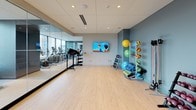 This contemporary fitness studio offers a thoughtfully designed, single-floor space that seamlessly blends functionality with a clean, modern aesthetic. The open floor plan is anchored by light hardwood-style flooring, which extends throughout the entire area and contributes to a bright, welcoming atmosphere.

A defining architectural feature is the expansive floor-to-ceiling glass mirror wall, which runs the length of the studio and is flanked by large windows offering sweeping city views. This combination of reflective surfaces and natural light makes the space feel exceptionally airy and open.

The cardio zone is clearly visible through the glass partition and houses a row of stationary bikes, treadmills, and other aerobic machines. The open studio area is designed for group fitness and personal training, equipped with a rack of dumbbells in varying weights, kettlebells, plyo boxes, medicine balls, stability balls, yoga mats, and resistance accessories. A wall-mounted ballet barre provides additional versatility for barre or stretch classes.

Multiple flat-screen televisions are mounted on the soft blue-gray accent walls, enhancing the workout experience. Recessed lighting and an integrated ceiling speaker system complete the polished interior. The overall ambiance is professional, energizing, and well-suited for a wide range of fitness activities.