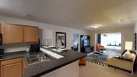 Located at 215 William Penn Plaza in Durham, NC, this single-floor residence offers a thoughtfully designed open-concept layout that seamlessly connects the kitchen, dining, and living areas. The kitchen is outfitted with dark granite countertops that extend into a breakfast bar, a double basin sink, and ample cabinetry, providing both functionality and a sleek aesthetic. The textured ceiling adds depth and character throughout the space. The open flow from the kitchen leads directly into the living area, which is furnished with a bold zebra-print area rug, a media console, and comfortable seating including a distinctive orange accent chair — creating a lively yet cohesive atmosphere. Natural light filters in through large windows, brightening the living space and adding warmth to the overall ambiance. A decorative framed mirror serves as a stylish focal point along the main corridor wall, enhancing the sense of openness. Beyond the living area, a bedroom is visible, offering a private retreat with its own subtle lighting. The single-floor plan ensures easy accessibility and a connected feel throughout, making this a well-balanced and functional living space suited for everyday comfort.