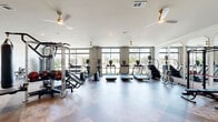 Located at 4850 Rouzan Square Ave in Baton Rouge, Louisiana, this well-appointed fitness center offers a generously sized, single-floor layout designed to accommodate a comprehensive workout experience. The space is organized into distinct zones, seamlessly flowing from one area to the next across a striking large-format checkerboard-patterned tile floor that adds visual depth and character throughout.

The main exercise area is lined with floor-to-ceiling windows that flood the space with abundant natural light and provide open views of the surrounding landscape. A row of treadmills and elliptical trainers runs along the window wall, complemented by a dedicated strength training section featuring cable machines, selectorized equipment, free weights, and kettlebells. A functional training zone includes a rower, medicine ball rack, stability balls, and resistance accessories.

Arched accent mirrors are positioned along one wall, enhancing the sense of openness while serving a practical purpose for form monitoring. Multiple modern ceiling fans with gold hardware circulate air efficiently throughout. A separate, more open room provides flexible space for stretching or group workouts, and a built-in bench with cubby storage near the entrance offers a convenient area to prepare before or unwind after a session. Sports-themed artwork and crown molding add polished, refined touches to the overall ambiance.