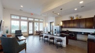 This single-floor amenity facility offers a thoughtfully designed collection of shared spaces built to support an active and convenient lifestyle. The centerpiece is a spacious open-plan communal kitchen and lounge area, featuring rich dark cabinetry, sleek black granite countertops, stainless steel appliances, and a large island with bar seating. Pendant lighting hangs overhead, while expansive windows and glass doors flood the space with natural light and provide views to the outdoors. An adjacent lounge area includes comfortable seating and a wall-mounted flat-screen TV, creating an inviting space for relaxation and socializing. Wood-look tile flooring runs throughout, lending warmth and continuity to the common areas. The fitness center is well-appointed across two connected rooms, offering cardio equipment including treadmills, a multi-station cable machine, free weights, and exercise balls, all set on patterned carpet with large mirrors and windows providing a bright workout environment. A dedicated business center provides built-in desk workstations, computers, and a printer for resident use. The laundry room is outfitted with multiple stackable and side-by-side washer and dryer units. A transitional corridor with dual water fountains and decorative tile accents connects the various amenity zones seamlessly.