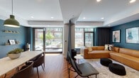 Located at 1330 Boylston St in Boston's vibrant Fenway neighborhood, this contemporary single-floor space offers a thoughtfully designed open-plan layout that seamlessly connects its living and dining areas. Rich hardwood flooring runs throughout, providing a warm foundation that contrasts beautifully with the deep teal accent walls and crisp white ceilings fitted with recessed lighting.

The living area is anchored by a generously sized tan leather sectional sofa, positioned beneath a trio of framed abstract artwork on a bold teal feature wall. Floor-to-ceiling glass doors and large windows flood the space with natural light and offer direct views of an outdoor patio and surrounding greenery, creating a strong indoor-outdoor connection.

The dining area features a marble-topped table with upholstered seating beneath striking green pendant lights, all set against the same deep teal wall that houses a wall-mounted flat-screen TV, floating media console, and decorative gold shelving. A dedicated lounge chair and side table complete the social arrangement.

The open floorplan and strategic use of a partial dividing column allow each zone to feel purposeful yet connected. The overall ambiance is modern and inviting, blending bold color choices with warm natural materials for a space that is both functional and aesthetically refined.