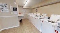 This shared laundry facility is designed to serve multiple residents efficiently, offering a clean and functional environment for everyday laundry needs. The room is lined along one wall with a row of at least eight numbered top-loading washing machines, each clearly labeled for easy identification. A matching set of dryers runs alongside, providing a seamless workflow from washing to drying. The machines are arranged in a single, organized row, maximizing the usable floor space within the room.

The space features neutral beige walls accented by white chair-rail molding, giving it a tidy, well-maintained appearance. Overhead fluorescent lighting ensures the room is brightly and evenly lit. The floor is covered in light-toned ceramic tile, which is easy to clean and contributes to the room's practical, no-fuss aesthetic.

A fold-down table is mounted near the entrance, providing a dedicated surface for sorting and folding laundry. A posted "Laundry Room Rules" sign and instructional notices are displayed on the walls, keeping users informed of proper usage guidelines. The overall layout is efficient and user-friendly, prioritizing functionality and cleanliness throughout.