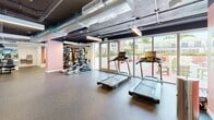 Located at 1470 NW 36th St in Miami, FL, this amenity space combines a well-appointed fitness center with a lively outdoor patio, delivering a complete wellness experience in a single level. The gym features an industrial-modern aesthetic, with exposed ductwork running across a dark ceiling, rubber-floored open areas, and full-length mirrors that enhance the sense of space. Equipment includes treadmills, elliptical machines, a cable strength station, a dumbbell rack, and open floor space for functional training. Large glass doors and floor-to-ceiling windows flood the interior with natural light and provide seamless visual and physical access to the outdoor patio. The patio area is boldly designed with a striking pink and light grey color-blocked floor, evoking Miami's vibrant energy. It features lounge chairs for sunbathing, lime-green Adirondack chairs, lush tropical plantings including palm trees, raised garden beds, and a casual game area. The surrounding multi-story building facade features bright lime-green balcony accents, reinforcing the playful, contemporary architectural identity. Together, the gym and patio offer a cohesive amenity environment suited to both active fitness routines and relaxed outdoor leisure in the heart of Miami.