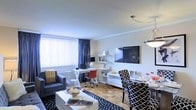 This single-floor space showcases a thoughtfully designed open-concept layout that combines living, dining, and work areas into one cohesive environment. The warm parquet hardwood flooring adds a classic touch that anchors the entire space, complementing the neutral white walls and creating a welcoming atmosphere.

The living area features a comfortable upholstered armchair paired with a sleek white coffee table atop a bold black-and-white patterned area rug, adding visual interest to the space. A large window dressed with white vertical blinds and navy blue curtains allows ample natural light to fill the room, enhancing its airy feel.

A wall-mounted flat-screen television serves as a focal point, flanked by a white shelving unit that provides both display and storage solutions. Adjacent to the living area, a dedicated workspace includes a white desk with a computer setup, offering a functional home office nook.

The dining area is furnished with a round table surrounded by stylish black-and-white patterned chairs, with a contemporary pendant light overhead completing the look. The overall ambiance is modern, clean, and versatile, making excellent use of the available space.