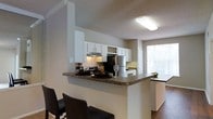 This single-floor residence offers a thoughtfully designed layout that emphasizes open-concept living. The kitchen serves as a central highlight, featuring crisp white cabinetry that extends to the ceiling, complete with a distinctive wine rack detail adding a refined touch. Dark granite-style countertops provide a striking contrast against the white finishes, and stainless steel appliances, including a refrigerator and range with an under-cabinet hood, equip the space for everyday functionality. A raised breakfast bar with seating extends from the kitchen peninsula, creating a natural gathering point that connects the kitchen to the adjacent living areas. The continuity of the open floor plan allows natural light to flow freely throughout, with a large window visible beyond the kitchen flooding the space with brightness. Hardwood-style flooring anchors the living areas, complementing the neutral wall tones and textured ceilings that run throughout. The overall ambiance is clean, warm, and inviting, blending practical design with comfortable aesthetics suited for both everyday living and casual entertaining.