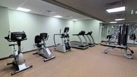 Located at 10 Landing Lane in New Brunswick, NJ, this fitness center offers a dedicated space for cardio exercise and general wellness. The room features a neutral color palette with light green walls and a wall-to-wall carpeted floor, creating a clean and comfortable environment for workouts. Recessed panel lighting in the drop ceiling ensures bright, even illumination throughout the space. The facility is equipped with a selection of True Fitness cardio machines, including multiple treadmills, an elliptical trainer, and a recumbent stationary bike, all positioned to allow ample room for movement between equipment. A floor-length mirror along one wall enhances the sense of space and allows users to monitor their form during exercise. Free weights are also available, visible along the perimeter of the room. The layout is practical and uncluttered, making efficient use of the available floor area. Ventilation is provided through ceiling-mounted air vents, contributing to a comfortable workout environment. Overall, the fitness center presents a functional and well-maintained amenity suited for everyday cardio training and light strength work.