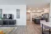 This thoughtfully designed single-floor residence offers a seamless open-concept layout connecting the living, dining, and kitchen areas, all unified by warm light wood flooring. The living area provides ample room for relaxed entertaining, featuring large windows dressed with black-and-white curtains that allow generous natural light to fill the space. A dedicated dining nook sits adjacent to the living area, anchored by a sleek white table and accented by a round gold-framed mirror and a window with outdoor views.

The kitchen is a standout feature, showcasing two-tone cabinetry with white upper cabinets and dark wood lower cabinets, a geometric tile backsplash, light quartz countertops, and a full suite of stainless steel appliances including a gas range, over-the-range microwave, side-by-side refrigerator, and dishwasher. A dark-toned kitchen island with bar seating adds both functionality and style.

The bedroom is a calm retreat with neutral tones, light wood flooring, and a bright window overlooking greenery. The full bathroom features a combination tub and shower with sliding glass doors, a wood-tone vanity with a light countertop, and a bold green accent wall that adds a distinctive touch to the clean, modern design.
