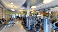 Located at 1000 Antietam Way in Fayetteville, North Carolina, this fitness center offers a thoughtfully designed, single-floor workout facility built to accommodate a wide range of exercise needs. The space features a generous open floor plan that houses an extensive selection of strength training machines, cable systems, and free weights, all arranged to allow comfortable movement between stations. Large pendant light fixtures and recessed lighting illuminate the entire area evenly, creating a bright and energizing atmosphere. Floor-to-ceiling mirrors line the walls, visually expanding the space while allowing users to monitor their form throughout their workouts. The interior design incorporates bold accent colors — including vibrant green and warm orange wall segments — paired with exposed ductwork overhead, lending the facility an industrial-modern aesthetic. The polished concrete flooring and rubberized workout zones provide both durability and functionality underfoot. Structural columns integrated throughout the layout help define workout zones without interrupting the open feel of the space. The overall ambiance is clean, modern, and motivating, making this fitness center a well-equipped destination for both strength and conditioning training.