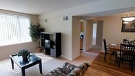 This single-floor residence offers a thoughtfully arranged open layout that promotes a natural flow between living spaces. The living room serves as the central hub of the home, featuring neutral beige walls and a combination of carpeted and hardwood flooring that adds warmth and visual variety to the space. Natural light filters through the windows, casting pleasant light across the room and highlighting the clean, minimalist design aesthetic.

Architectural archways connect the living area to the rest of the home, providing an open and airy feel while maintaining a sense of defined spaces. Through these openings, the kitchen is visible, featuring light wood cabinetry and ample counter space, providing a functional and inviting cooking environment. A hallway extends further beyond, leading to additional rooms within the residence.

The living room itself is generously proportioned, offering comfortable space for both relaxing and entertaining. Ceiling-mounted HVAC vents throughout the space ensure climate control across the entire floor. The neutral color palette of the walls and flooring creates a versatile backdrop, making the space easy to personalize and furnish. Overall, this single-floor home balances practicality with a welcoming ambiance, making it a comfortable and functional living environment.