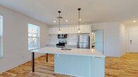 This single-floor residence showcases a clean, contemporary design highlighted by light hardwood flooring and crisp white walls throughout the main living areas. The open-concept kitchen is a standout feature, centered around a large island with a white quartz countertop, an undermount sink, and pendant lighting overhead. Cabinetry is finished in a two-tone style, with white upper cabinets and gray lower cabinets, complemented by a classic white subway tile backsplash and stainless steel appliances including a gas range, microwave, and refrigerator. The kitchen flows seamlessly into the living area, which is anchored by a sleek wall-mounted fireplace, creating a warm and inviting focal point. Multiple windows throughout the open-plan space invite abundant natural light. The bedrooms feature soft neutral carpeting, ceiling fans, and windows with blinds that offer a comfortable and restful atmosphere. The bathrooms are finished with care, featuring penny-tile flooring, a floating vanity with a framed mirror, and a walk-in shower enclosed by a bold black-framed glass panel door set against all-white tile walls. The overall ambiance is bright, modern, and move-in ready.