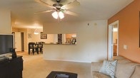 This single-floor residence is designed around an open-concept floor plan that seamlessly connects the living, dining, and kitchen areas into one cohesive space. The living area is anchored by a ceiling fan with an integrated light fixture, providing both airflow and ambient lighting. Neutral-toned carpet runs throughout, contributing to a warm and comfortable atmosphere.

A half-wall with a pass-through counter serves as the dividing element between the living space and the kitchen, maintaining visual openness while subtly defining each zone. The kitchen features oak-toned cabinetry and a range hood, with the pass-through offering a convenient serving or interaction point between rooms.

Adjacent to the kitchen, the dining area accommodates a full dining table and chair set beneath a pendant light, creating a dedicated space for meals. Framed artwork on the walls adds a touch of personality to the dining zone.

The overall palette is light and neutral, with cream-colored walls and warm wood accents throughout. The layout prioritizes a sense of openness and flow, making the space feel connected and easy to navigate across all living areas.