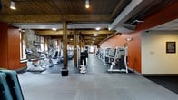 This fitness facility showcases a compelling blend of industrial and rustic architectural character. The space is defined by its exposed heavy-timber wooden columns and ceiling beams, which contrast beautifully with the bold terracotta-orange accent walls and original brick detailing along the perimeter. Overhead, industrial-style lighting fixtures are suspended from the wood-planked ceiling, casting even illumination throughout the floor.

The single-story layout is elongated and generously proportioned, offering ample room for a wide variety of exercise equipment arranged in organized rows. Along one side, a lineup of cardio machines—including ellipticals, stationary bikes, and treadmills—provides dedicated aerobic training space. Opposite, a row of strength and resistance machines lines the vivid orange wall, offering a comprehensive selection for targeted muscle training.

The flooring transitions from rubberized gym matting in the equipment zones to a carpet-like surface in open areas, supporting both functionality and comfort. Exposed ductwork and utility conduits run along the ceiling, reinforcing the industrial aesthetic. The overall ambiance is energetic yet grounded, combining the warmth of natural wood with the practicality of a well-equipped fitness environment.