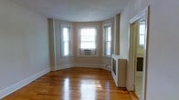 This single-floor residence offers a blend of classic architectural character and functional living. The standout feature is a graceful bay window arrangement, with three large windows curving outward to flood the interior with abundant natural light. The windows are fitted with white trim that complements the neutral, light-toned walls throughout the space.

The flooring is a highlight of the home — rich, warm-toned hardwood runs throughout, exhibiting a natural luster that reflects the incoming daylight and adds a sense of depth and warmth to the rooms. Classic baseboard trim lines the perimeter of the walls, reinforcing the traditional aesthetic of the space.

A built-in radiator unit is positioned along one wall, indicative of a well-established heating system consistent with classic residential construction. An additional doorway visible from the main room suggests a connected, flowing layout that provides easy access between living areas.

Overall, the space carries a timeless residential character — bright, clean, and well-proportioned, with architectural details that speak to its classic construction. The combination of natural light, hardwood floors, and bay window geometry makes this a notably inviting and functional single-floor living space.
