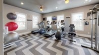 This fitness center is a thoughtfully designed single-level space that balances functionality with modern aesthetics. The room is immediately distinguished by its bold chevron-patterned carpet in shades of taupe, brown, and cream, which runs throughout the entire floor area and adds a striking visual element to the space.

The cardio zone is positioned along a windowed wall, where multiple treadmills and elliptical machines are arranged in a row. Natural light filters in through several white-framed windows fitted with blinds, while wall-mounted flat-screen televisions are placed at eye level for entertainment during workouts. A ceiling fan with integrated lighting provides additional airflow and illumination.

A dedicated strength training area features a multi-function cable machine, a weight bench, and a tiered dumbbell rack. Large mirrors line one wall, visually expanding the space and allowing users to monitor their form. Exercise balls and additional accessories are also available.

The neutral taupe walls, white trim, and recessed lighting contribute to a clean, contemporary atmosphere. An exit door and a bulletin board are conveniently located near the entrance, and a water fountain is accessible within the space. The overall layout is open and organized, supporting a variety of workout routines.