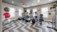 This fitness center is a thoughtfully designed single-level space that balances functionality with modern aesthetics. The room is immediately distinguished by its bold chevron-patterned carpet in shades of taupe, brown, and cream, which runs throughout the entire floor area and adds a striking visual element to the space.

The cardio zone is positioned along a windowed wall, where multiple treadmills and elliptical machines are arranged in a row. Natural light filters in through several white-framed windows fitted with blinds, while wall-mounted flat-screen televisions are placed at eye level for entertainment during workouts. A ceiling fan with integrated lighting provides additional airflow and illumination.

A dedicated strength training area features a multi-function cable machine, a weight bench, and a tiered dumbbell rack. Large mirrors line one wall, visually expanding the space and allowing users to monitor their form. Exercise balls and additional accessories are also available.

The neutral taupe walls, white trim, and recessed lighting contribute to a clean, contemporary atmosphere. An exit door and a bulletin board are conveniently located near the entrance, and a water fountain is accessible within the space. The overall layout is open and organized, supporting a variety of workout routines.