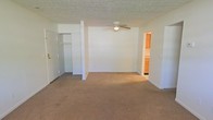This single-floor space centers around a generously sized main room finished with neutral wall-to-wall carpeting that lends warmth underfoot. A ceiling fan with an integrated light fixture is mounted overhead, providing both air circulation and ambient lighting. The textured ceiling adds a classic touch throughout. An open closet along one wall offers convenient storage, complete with a hanging rod and visible shelving space. The room connects directly to a kitchen, visible through a wide doorway, which features warm wood-toned cabinetry that adds a natural contrast to the otherwise neutral color palette. White walls and understated finishes throughout create a clean, blank-canvas atmosphere well suited for personalization. The single-floor layout ensures easy accessibility and a straightforward flow between spaces.