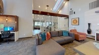 Located at 9999 E Yale Ave in Denver, Colorado, this single-floor community clubhouse offers a thoughtfully designed gathering space that blends contemporary style with inviting comfort. The open-plan layout centers around a spacious lounge area anchored by a large sectional sofa, accent armchairs, and a wall-mounted flat-screen TV positioned above a rich wood media console — all beneath soaring vaulted ceilings with exposed wood beams and a skylight that floods the space with natural light. A decorative geometric lattice ceiling panel serves as a dramatic focal point above the kitchenette, which features a long white quartz island with bar seating, dark cabinetry, a utility sink, and pendant globe lighting. Adjacent to the kitchenette, a casual high-top bistro table with stools is positioned beside tall windows overlooking lush greenery, providing an airy nook ideal for informal gatherings. A dedicated business center area with computer workstations and a printer offers functional workspace within the communal setting. Throughout the space, curated décor including sculptural wall art, driftwood pieces, and bold accent pillows in teal and orange contribute to a warm yet modern aesthetic. Large windows on multiple walls invite abundant natural light and connect the interior to the surrounding outdoor landscape.