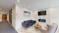 Located at 205 S Westmore Ave in Lombard, Illinois, this sophisticated lobby offers a refined and welcoming first impression across a single well-appointed floor. The space is divided into a hallway, a lounge-style living area, and an additional glass-enclosed room, each reflecting a high standard of interior design.

The hallway entrance greets visitors with marble-look porcelain tile flooring accented by gold veining, transitioning into a patterned carpet runner. A striking sputnik-style chandelier illuminates the entry, complemented by recessed lighting and elegant wainscoting that runs throughout. Large black-framed glass doors and floor-to-ceiling windows contribute a modern architectural contrast to the classic millwork.

The lounge area serves as the centerpiece of the lobby, featuring a plush navy velvet sofa, contemporary accent chairs, and a round wood coffee table. A dramatic marble-look accent wall houses a sleek linear electric fireplace and a wall-mounted flat-screen television above it, creating a focal point that blends warmth with modern style. Crown molding and panel wainscoting frame the crisp white walls throughout.

The glass-enclosed room, accessible via a set of steel-framed glass doors, offers light wood flooring and a clean, open layout with recessed lighting — ideal for a flex-use or private meeting space. Together, these areas form a polished, upscale lobby environment.