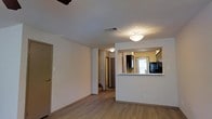 This two-floor space offers a practical and functional layout with a clean, neutral aesthetic. The main living area features light wood-style flooring and white walls, creating a bright and open atmosphere. A textured ceiling with flush-mount lighting adds a classic touch throughout the space.

A defining feature is the pass-through kitchen opening, which connects the kitchen to the living area. This opening is framed by a countertop bar ledge, providing a convenient serving or casual dining surface. Within the kitchen, dark cabinetry contrasts against light walls, and a tile backsplash adds visual interest. A window allows natural light to filter into the kitchen area, and black appliances complete the space.

The living area flows naturally toward a hallway that leads further into the unit, providing access to additional rooms and the second floor. The overall layout is straightforward and efficient, with neutral finishes throughout that offer a versatile backdrop. The combination of light flooring, white walls, and connected living and kitchen spaces gives the home a cohesive and welcoming feel.