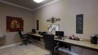 This single-floor business center offers a functional and well-appointed workspace designed to accommodate multiple users simultaneously. The room features a continuous granite-topped counter running along the walls, providing ample desk space for several workstations equipped with desktop computers and a shared printer. Comfortable ergonomic office chairs are positioned at each station, ensuring a practical setup for extended use. The space is finished with warm neutral-toned walls accented by crown molding, adding a polished architectural detail that elevates the overall aesthetic. Decorative wall art, including an ornate metal wall piece and framed panels, contribute a touch of personality to the functional environment. A cork bulletin board mounted on the wall serves as an informational display area. Light-toned tile flooring runs throughout, complementing the room's neutral color palette. Overhead flush-mount lighting provides consistent illumination across the workspace. The overall ambiance balances practicality with a welcoming, professionally finished atmosphere suited for business or administrative tasks.
