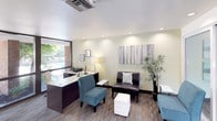 This single-floor office space is thoughtfully designed to serve as a professional leasing or property management environment. The interior features a cohesive aesthetic throughout, with soft sage-green walls, recessed lighting, and patterned carpeting that together create a calm, polished atmosphere.

The reception area greets visitors with a mix of seating options, including tufted leather sofas, teal accent chairs, and white upholstered ottomans, complemented by tall decorative bamboo plants and contemporary wall art. A front desk with a sleek, low-profile design provides a functional workspace while maintaining an open, inviting feel.

A private office off the main area is equipped with an L-shaped white and dark wood desk, dual monitors, and additional seating for consultations, making it well-suited for one-on-one meetings.

Large floor-to-ceiling windows along one wall flood the space with natural light while offering direct views of a landscaped outdoor courtyard with a swimming pool, mature trees, and brick architectural detailing. This visual connection to the outdoors enhances the overall ambiance, lending the interior a bright and open quality. The space is efficiently organized and professionally appointed throughout.