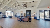 This expansive single-floor clubhouse or community lounge offers a generously sized, open-plan interior designed with a contemporary aesthetic. Soaring vaulted ceilings with exposed beam detailing create a dramatic sense of height and volume throughout the main gathering area. Light hardwood flooring flows across much of the space, complemented by sections of decorative geometric tile that define distinct seating zones.

The main lounge features multiple seating arrangements, including plush blue sofas, warm-toned orange accent chairs, and leather-upholstered furniture grouped around a striking floor-to-ceiling black tile fireplace wall, complete with a mounted flat-screen television and a linear gas fireplace below. A billiards table anchors a recreational zone nearby.

A dedicated kitchenette area showcases slate-blue cabinetry with gold hardware and a dramatic veined white quartz countertop, accented by a playful neon sign. Adjacent to this is a café-style workspace with a marble-topped island and bar seating beneath expansive floor-to-ceiling windows, flooding the area with natural light and offering outdoor views. A semi-private meeting room with glass partition walls provides a quieter enclave. Woven pendant lights and sleek track lighting complete the polished, inviting atmosphere.