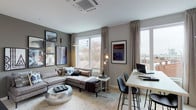 This thoughtfully designed single-floor residence showcases a contemporary aesthetic throughout, combining functional layout with modern finishes. The open-plan living and dining area serves as the heart of the unit, featuring warm neutral-toned walls, light hardwood-style flooring, and large windows that frame rooftop cityscape views, flooding the space with natural light. A generously sized sectional sofa anchors the living zone, complemented by a curated gallery wall and a reflective metallic accent table. A sleek bar-height dining table with modern chairs doubles as a workspace, offering versatility. The kitchen runs along a galley-style layout, featuring two-tone cabinetry with dark lower cabinets and crisp white upper cabinets, white quartz countertops, a light grey tile backsplash, stainless steel appliances, and open industrial-style shelving. The bedroom offers a calm, neutral retreat with soft lighting and direct access to a spacious walk-in closet equipped with wire shelving and hanging storage. The full bathroom features large-format dark tile flooring, a combined shower and soaking tub with a patterned curtain, a white vanity countertop, and grey cabinetry, completing the cohesive modern palette found throughout the residence.
