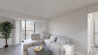 This single-floor unit offers a well-organized layout across three bedrooms, one bathroom, a combined living and dining area, and a galley-style kitchen. The living room is the focal point of the unit, featuring large floor-to-ceiling windows and a sliding glass door that opens onto a balcony with outdoor views of surrounding greenery. Light-toned laminate flooring and a neutral white and grey palette carry throughout the space, creating a clean, cohesive aesthetic.

The galley kitchen is efficiently designed with white cabinetry, quartz countertops, subway tile backsplash, stainless steel appliances including a refrigerator and dishwasher, and a stainless undermount sink — all contributing to a bright, modern feel. Adjacent to the kitchen, the dining area accommodates a round table beneath a pendant light fixture.

The primary bedroom is the most spacious of the three, while the additional bedrooms are compact and functional, each with laminate flooring and a window. The bathroom features dark slate-style floor tiles, a white vanity with under-cabinet storage, a wall mirror, and a standing shower, finished in a clean, minimal style throughout.