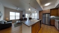 This single-floor residence offers a thoughtfully designed open-concept layout that seamlessly connects the kitchen, dining, and living areas. The kitchen serves as the centerpiece of the space, anchored by a generously sized island with a dark countertop and an integrated double sink. Warm wood cabinetry lines the kitchen walls, complemented by stainless steel appliances including a full-size refrigerator and dishwasher. Pendant lighting hangs above the island, adding a modern touch to the functional workspace. Hardwood-style flooring runs throughout, lending warmth and continuity across the entire floor plan. The open dining area transitions naturally into the living room, which is furnished with comfortable seating arranged around a low coffee table atop a striped area rug. A ceiling fan adds both practicality and a casual aesthetic to the living space. Multiple large windows flood the interior with abundant natural light, offering views to the outdoors and creating an airy, welcoming ambiance. The layout is efficient yet spacious, making excellent use of the available footprint while maintaining a clean, contemporary feel throughout.