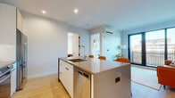 This single-floor space showcases a thoughtfully designed modern interior with a cohesive aesthetic carried throughout each room. Light hardwood flooring runs continuously across the main areas, complementing the clean white walls and creating a bright, airy atmosphere.

The primary kitchen is equipped with flat-front white cabinetry, a marble-effect backsplash, stainless steel appliances including a refrigerator, range, and microwave, and a substantial island with a dark quartz countertop and integrated sink. A second kitchen features a similarly styled island with a stainless steel countertop, dishwasher, and recessed lighting overhead.

The living area benefits from floor-to-ceiling windows and glass door access to an outdoor space, flooding the room with natural light. A wall-mounted mini-split unit provides climate control.

The bedroom offers a calm, restful setting with soft neutral tones, recessed lighting, and a spacious closet. Two bathrooms are finished with large-format stone-look tile flooring, white subway tile walls, floating wood vanities, and mirrored medicine cabinets. One bathroom includes a glass-enclosed walk-in shower with a rainfall showerhead.