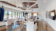 This thoughtfully designed single-floor gathering space blends coastal-inspired aesthetics with modern architectural detail. The open-concept layout is anchored by a striking coffered ceiling with warm wood-planked insets, supported by crisp white structural beams and accented by a mix of globe and industrial-style pendant lighting. Underfoot, richly marbled epoxy floors in swirling tones of copper, blue, and grey reflect light throughout the space, adding a dynamic visual element that complements the clean white walls.

The main lounge area is furnished with plush white slipcovered sofas and armchairs arranged over a bold geometric area rug, creating an inviting social setting. Multiple dining tables with slipcovered chairs are positioned near expansive floor-to-ceiling sliding glass doors, which open directly to an outdoor pool and patio area, flooding the interior with natural light and views of the surrounding landscape. Royal blue drapery provides a rich color accent against the bright white walls.

A well-appointed kitchen area features white shaker cabinetry, a farmhouse sink, dark countertops, stainless steel appliances, open floating shelves, and a warm wood-tone accent wall. A large island with bar seating adds functional versatility. Bold abstract artwork is displayed throughout, contributing to the space's vibrant and contemporary character.