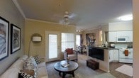 This single-story space showcases a thoughtfully designed open-concept living and dining area finished with warm neutral tones throughout. The living room is anchored by plush seating arrangements, including a sofa and accent chair adorned with patterned throw pillows, centered around a rustic round coffee table. Soft carpeting underfoot adds warmth and comfort to the space. Crown molding runs along the ceiling perimeter, lending an elevated, refined aesthetic, while a ceiling fan with an integrated light fixture provides both air circulation and ambient lighting. A track lighting system along the ceiling offers additional illumination, particularly toward the dining area. Natural light flows generously into the space through a full-length glass-paneled door flanked by large windows dressed with vertical blinds, providing both brightness and outdoor access. The dining area is visible in the background, featuring a small table and chairs, seamlessly connected to the living space. A large flat-screen television serves as a focal point along one wall, complemented by a dark wood media console. The overall ambiance is cozy and well-appointed, balancing functional living with comfortable, everyday design.