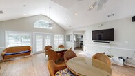 Located in Savannah, GA, this single-story clubhouse offers a welcoming and airy gathering space designed for community use. The main room is defined by striking vaulted ceilings that soar overhead, creating a sense of openness and volume. A signature arched transom window crowns the front wall, flooding the interior with natural light alongside multiple sets of white-shuttered French doors that open to the outdoors, offering views of the surrounding grounds and pool area.

The interior is finished with light hardwood flooring and neutral gray-toned walls accented by classic chair-rail wainscoting, lending the space a clean, coastal character. Ceiling fans are strategically positioned throughout to ensure comfort. The main gathering area is furnished with multiple wicker seating arrangements — including loveseats and lounge chairs with bold blue cushions — as well as round glass-top dining tables with wicker chairs, accommodating both casual socializing and small group dining.

A wall-mounted flat-screen television with a dedicated media shelf provides entertainment options for residents. A pass-through opening connects the main room to a functional kitchenette equipped with cabinetry, a microwave, and a range, supporting events and gatherings. Multiple French door entries throughout the space ensure easy access from various points, making this clubhouse a versatile and inviting hub for the community.