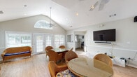 Located in Savannah, GA, this single-story clubhouse offers a welcoming and airy gathering space designed for community use. The main room is defined by striking vaulted ceilings that soar overhead, creating a sense of openness and volume. A signature arched transom window crowns the front wall, flooding the interior with natural light alongside multiple sets of white-shuttered French doors that open to the outdoors, offering views of the surrounding grounds and pool area.

The interior is finished with light hardwood flooring and neutral gray-toned walls accented by classic chair-rail wainscoting, lending the space a clean, coastal character. Ceiling fans are strategically positioned throughout to ensure comfort. The main gathering area is furnished with multiple wicker seating arrangements — including loveseats and lounge chairs with bold blue cushions — as well as round glass-top dining tables with wicker chairs, accommodating both casual socializing and small group dining.

A wall-mounted flat-screen television with a dedicated media shelf provides entertainment options for residents. A pass-through opening connects the main room to a functional kitchenette equipped with cabinetry, a microwave, and a range, supporting events and gatherings. Multiple French door entries throughout the space ensure easy access from various points, making this clubhouse a versatile and inviting hub for the community.