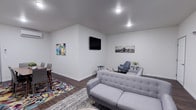 This single-floor space offers a clean, modern aesthetic defined by bright white walls, recessed lighting, and dark hardwood-style flooring that runs throughout. The open-concept layout seamlessly connects the living and dining areas, creating a cohesive and functional environment. The living area is anchored by a tufted gray sofa and complemented by a contemporary blue accent chair, a small side table with a potted plant, and a wall-mounted flat-screen television. Two distinct area rugs — one featuring a bold geometric multicolor pattern and another in a neutral tone — help define separate zones within the open space. Colorful abstract wall art adds a vibrant touch, while a black-and-white cityscape print provides a contrasting, urban-inspired accent. The dining area, positioned to one side, features a high-top table with matching chairs, ideal for casual dining or work. Recessed ceiling lights provide consistent, even illumination throughout the space. The overall ambiance is contemporary and inviting, with a thoughtful balance of color, texture, and functionality that makes the space well-suited for both relaxing and gathering.