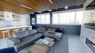 This single-floor space showcases a refined, modern design that blends industrial and contemporary elements with great cohesion. The most striking architectural feature is the slatted wood ceiling, which adds warmth and texture while contrasting handsomely against the bold navy blue walls. Integrated linear lighting runs through the ceiling slats, providing a clean, ambient glow throughout the space.

The main living area is anchored by a large, deep charcoal leather sectional sofa paired with a matching chaise lounge, both arranged around a two-tiered coffee table that combines a metal top surface with a natural wood lower shelf. A pool table positioned near the windows serves as both a functional amenity and a design focal point, reinforcing the space's social and recreational character.

A bank of large windows lines the far wall, flooding the interior with natural light and framing views of the surrounding urban skyline. Adjacent to the main lounge area, a compact kitchen counter with bar-height stools is visible, featuring a clean white countertop and pendant lighting above.

The overall ambiance is modern, sophisticated, and social — well-suited for both relaxation and entertainment.