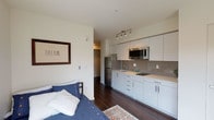 Located on the fourth floor at 5001 Brooklyn Avenue NE in Seattle's vibrant University District, Unit 406 is a thoughtfully designed studio that maximizes functionality within a well-organized open floor plan. Rich, dark hardwood flooring flows throughout the main living area, creating a warm contrast against the bright white walls and modern finishes.

The kitchen is efficiently laid out along one wall, featuring sleek white cabinetry — both upper and lower — stainless steel appliances including a refrigerator and microwave, a single-basin sink, and a gray tile backsplash that adds a contemporary touch. A dedicated closet with a rod sits adjacent to the kitchen, offering convenient storage.

The living and sleeping area is anchored by a large window that frames views of the surrounding neighborhood, allowing natural light to fill the space. A secondary window offers additional light and an elevated perspective of nearby rooftops and streetscapes.

The bathroom is clean and bright, featuring a full bathtub with shower, a frosted privacy window that brings in diffused natural light, and a simple, functional layout with light-toned flooring. The overall design of the unit is modern and cohesive, with a neutral palette that creates a calm, livable atmosphere suited for everyday urban living.