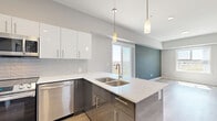Located at 425 Ballantrae Dr in Winnipeg, this well-designed one-bedroom, one-bathroom unit offers a thoughtful layout with modern finishes across a single floor. The contemporary kitchen is a standout feature, boasting two-tone cabinetry with high-gloss white upper cabinets and slate-grey lower cabinets, a classic subway tile backsplash, crisp white countertops, and a full suite of stainless steel appliances including a range, built-in microwave, and dishwasher. A peninsula with a double undermount sink and pendant lighting above adds both function and style, opening toward the living area and a sliding door that leads to a balcony with outdoor views. Light wood-look flooring flows through the main living areas, complementing the neutral grey and teal accent wall tones found throughout the unit. The bedroom is a comfortable size, bathed in natural light from a large window overlooking the surrounding neighborhood. It features plush carpeting, a soothing teal accent wall, and a closet fitted with a rustic wood-and-frosted-glass sliding door. The bathroom includes a full soaker tub with a clean white surround. An in-unit stacked washer and dryer adds everyday convenience to this move-in-ready space.