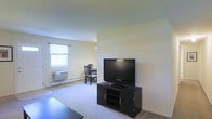 This single-floor space presents an open-concept layout that connects the living area to a dining space, creating a sense of flow and continuity throughout. The living area features wall-to-wall carpeting in a neutral tone, contributing to a quiet and comfortable atmosphere. A flat-screen television is positioned on a dark media console, serving as the focal point of the room. The walls are finished in a light, creamy hue that reflects natural light effectively. A window fitted with a simple curtain rod allows daylight to filter into the dining area, brightening the space and providing ventilation. A wall-mounted air conditioning unit and a baseboard heater are visible along the exterior wall, indicating year-round climate control options. The dining area accommodates a modest table and chairs, suitable for everyday use. Overhead, a simple light fixture provides additional illumination. The overall design is understated and functional, with a clean, minimalist aesthetic that provides a blank canvas for personalization.