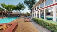 Located at 6700 Sandshell Blvd in Fort Worth, Texas, this outdoor swimming pool area offers a well-appointed recreational retreat designed for relaxation and leisure. The centerpiece is a generously sized swimming pool with clear, vivid turquoise water, framed by a textured aggregate concrete deck that surrounds the pool on multiple sides.

The pool deck is furnished with rows of cushioned chaise lounge chairs, providing ample space for sunbathing under the Texas sky. A covered pergola structure anchors one corner of the deck and houses a dedicated grilling and outdoor kitchen area, complete with additional seating and a patio umbrella — ideal for outdoor gatherings. Manicured hedges and mature shade trees are scattered throughout the space, adding natural greenery and providing welcome relief from the sun.

Adjacent to the pool stands a single-story clubhouse building with a distinctive red and stone exterior and a pitched shingle roof with a chimney. The building features an expansive wall of large-pane windows that overlook the pool area, allowing natural light to flow in while maintaining a visual connection to the outdoor space. The overall ambiance is vibrant and welcoming, combining functional amenities with attractive landscaping to create an inviting community outdoor environment.