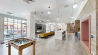Located at 1100 East Apache Boulevard in Tempe, Arizona, this expansive single-floor community space offers a generously sized, open-concept layout that seamlessly blends living, dining, and kitchen areas into one cohesive environment. The interior is defined by high ceilings adorned with cascading globe pendant lights in brushed brass, large-format tile flooring, and an abundance of natural light streaming through floor-to-ceiling windows and glass panel garage doors that open directly to an outdoor courtyard. The living area anchors the space with a bold mustard-yellow sectional sofa, a geometric patterned area rug, and a sleek gas fireplace set against a textured gray tile wall with a mounted flat-screen TV. A long white kitchen island with bar seating serves as both a functional and social hub. The space is enriched by vibrant, eclectic décor including colorful acrylic wall art, coral and white two-tone painted accent walls, tropical-print area rugs, and rich wood-paneled feature walls. A distinctive geometric laser-cut screen divider adds architectural interest. Recreational amenities include a foosball table and shuffleboard, while a glass-enclosed lounge area with colorful furnishings provides a lively secondary gathering spot. Tropical plants throughout add a fresh, organic element to the modern aesthetic.