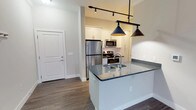 Located at 1221 E Broadway in Louisville, KY, this single-floor unit offers a thoughtfully designed layout with modern finishes throughout. The open-concept kitchen serves as the heart of the space, showcasing white shaker cabinetry, a subway tile backsplash, stainless steel appliances including a refrigerator, electric range, and microwave, and a center island with dark granite-style countertops and an undermount sink. Stylish pendant lights hang above the island, complemented by track lighting overhead, creating a well-lit and functional cooking and entertaining area. Rich wood-look flooring extends throughout the main living areas, adding warmth and continuity. The kitchen connects seamlessly to the living area, which features french doors that invite natural light and provide access to an outdoor space. A dedicated laundry area is conveniently tucked just off the kitchen. The bathroom is elegantly appointed with large-format marble-look tile in the walk-in shower, a white vanity with stone countertops, and matching wood-look flooring. The bedroom is generously proportioned, offering plush carpet, a ceiling fan, and two windows that fill the room with natural light, creating a comfortable and serene retreat.